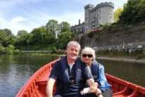 Smiling couple in lifejackets rowing a red boat on river with castle and green trees behind them (no visible text)