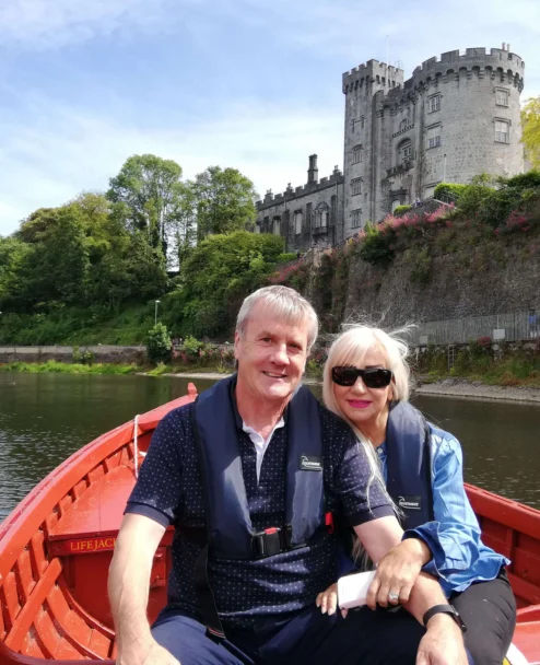 Smiling couple in lifejackets rowing a red boat on river with castle and green trees behind them (no visible text)