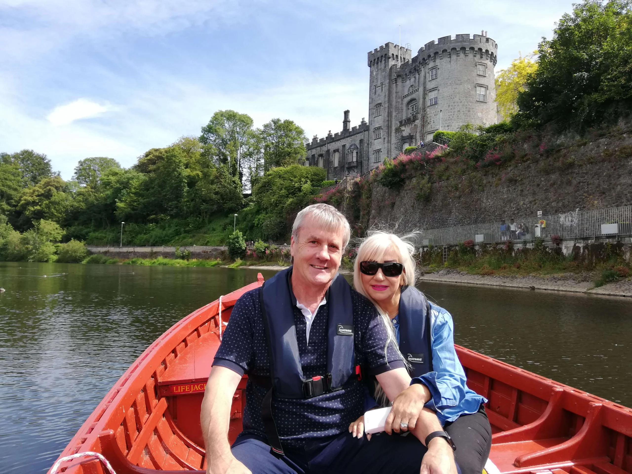 Smiling couple in lifejackets rowing a red boat on river with castle and green trees behind them (no visible text)