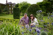 Smiling couple exploring a manicured hotel garden with trimmed hedges and purple globe thistles