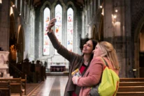 Two happy female friends taking a selfie inside a historic church with stained glass windows, holding a guidebook ALT_TEXT_TRANSCRIPTION: visible guidebook text "KILKENNY