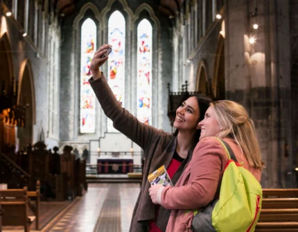Two happy female friends taking a selfie inside a historic church with stained glass windows, holding a guidebook ALT_TEXT_TRANSCRIPTION: visible guidebook text "KILKENNY