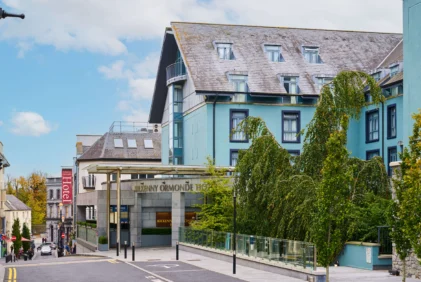 Family strolling past a welcoming coastal hotel entrance, blue façade and leafy trees, sign reads "Kilkenny Ormonde" (85 characters)