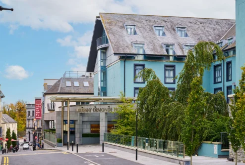 Family strolling past a welcoming coastal hotel entrance, blue façade and leafy trees, sign reads "Kilkenny Ormonde" (85 characters)