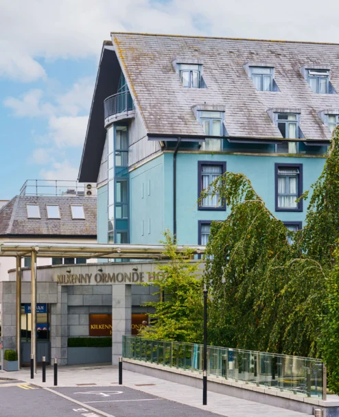 Family strolling past a welcoming coastal hotel entrance, blue façade and leafy trees, sign reads "Kilkenny Ormonde" (85 characters)