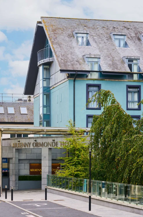 Family strolling past a welcoming coastal hotel entrance, blue façade and leafy trees, sign reads "Kilkenny Ormonde" (85 characters)