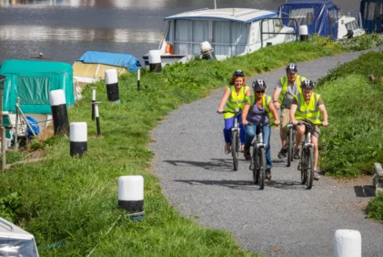 Family cycling along a waterside path by moored boats, wearing hi‑vis vests and helmets
