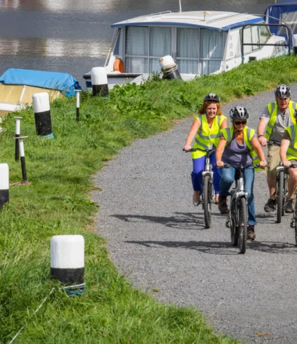 Family cycling along a waterside path by moored boats, wearing hi‑vis vests and helmets