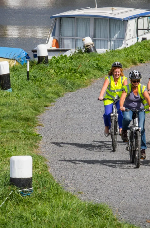Family cycling along a waterside path by moored boats, wearing hi‑vis vests and helmets