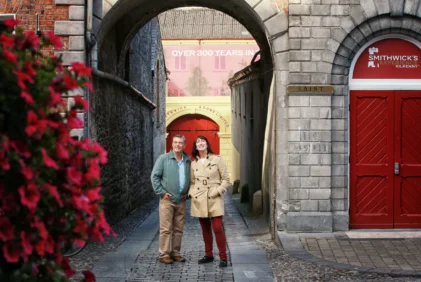 Mature couple strolling under a stone archway by Smithwick’s red doors, smiling together