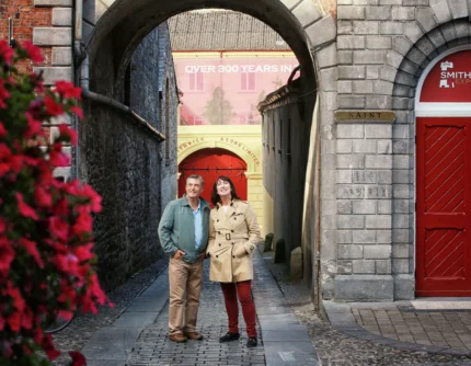 Mature couple strolling under a stone archway by Smithwick’s red doors, smiling together