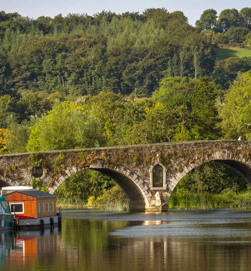 Family enjoying riverside boats by a historic stone bridge and leafy green hillside