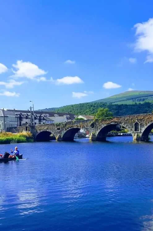 Family boating on a calm river near a historic stone bridge and riverside village under blue skies