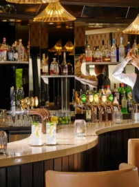 Friendly bartender shaking a cocktail behind a warm hotel bar with bottles and brass taps, family welcome