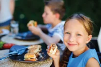 Smiling young girl eating a burger at an outdoor family meal with siblings nearby, sunny garden