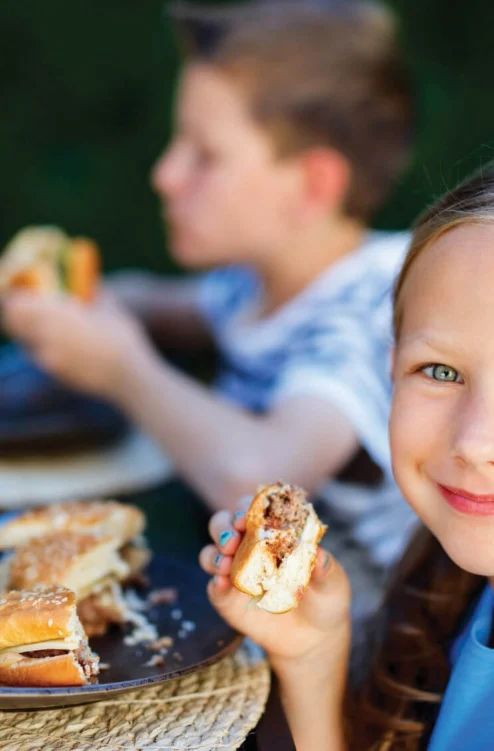 Smiling young girl eating a burger at an outdoor family meal with siblings nearby, sunny garden