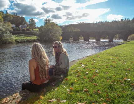 Two friends sitting by a sunlit riverbank near a stone arched bridge, relaxing together (no visible text)