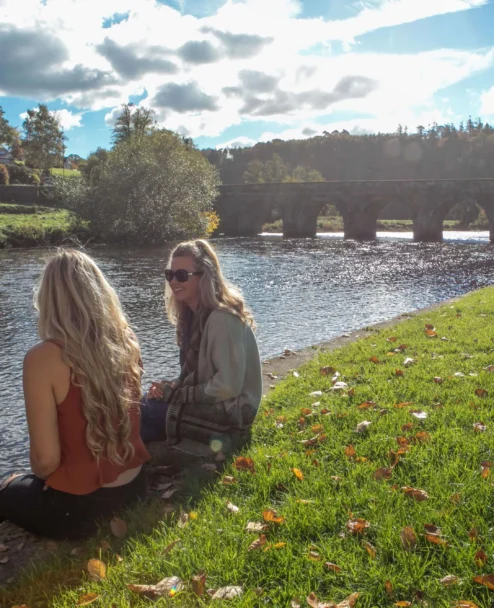 Two friends sitting by a sunlit riverbank near a stone arched bridge, relaxing together (no visible text)