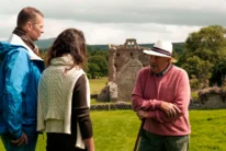 Couple with local guide exploring castle ruins on a green hillside, friendly conversation