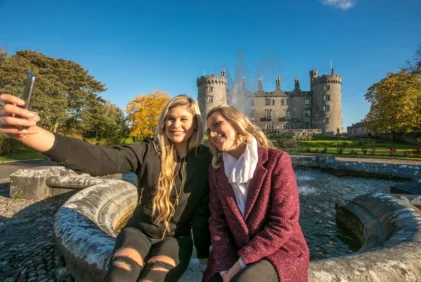 Mother and adult daughter smiling and taking a selfie by a fountain with a historic castle and gardens in the background, autumn light