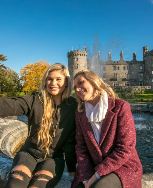 Mother and adult daughter smiling and taking a selfie by a fountain with a historic castle and gardens in the background, autumn light