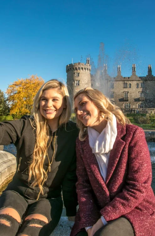 Mother and adult daughter smiling and taking a selfie by a fountain with a historic castle and gardens in the background, autumn light
