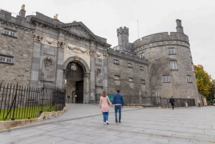 Couple holding hands stroll towards historic stone castle gatehouse on grey paved plaza, autumn trees visible