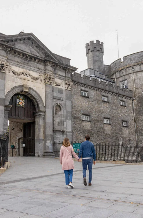 Couple holding hands stroll towards historic stone castle gatehouse on grey paved plaza, autumn trees visible