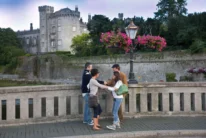 Family of four greeting on a riverside bridge with hanging pink flowers and historic castle backdrop