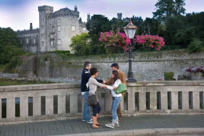 Family of four greeting on a riverside bridge with hanging pink flowers and historic castle backdrop