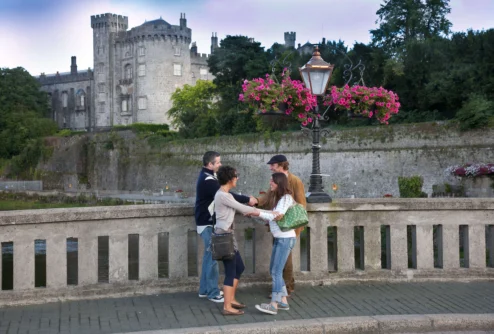 Family of four greeting on a riverside bridge with hanging pink flowers and historic castle backdrop