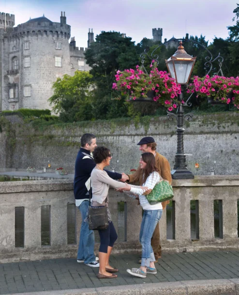 Family of four greeting on a riverside bridge with hanging pink flowers and historic castle backdrop