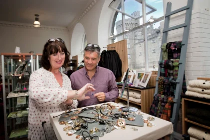 Couple browsing handmade jewellery at a cosy indoor gift shop with castle view through arched window