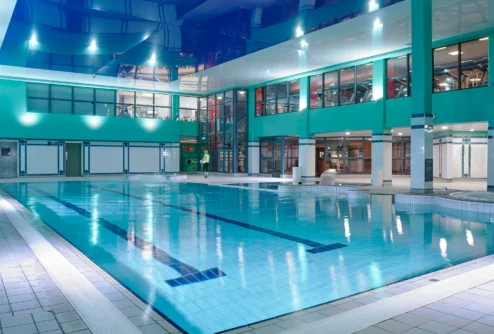 Family enjoying indoor hotel pool area with a parent and child walking by the water, turquoise décor (no visible text)