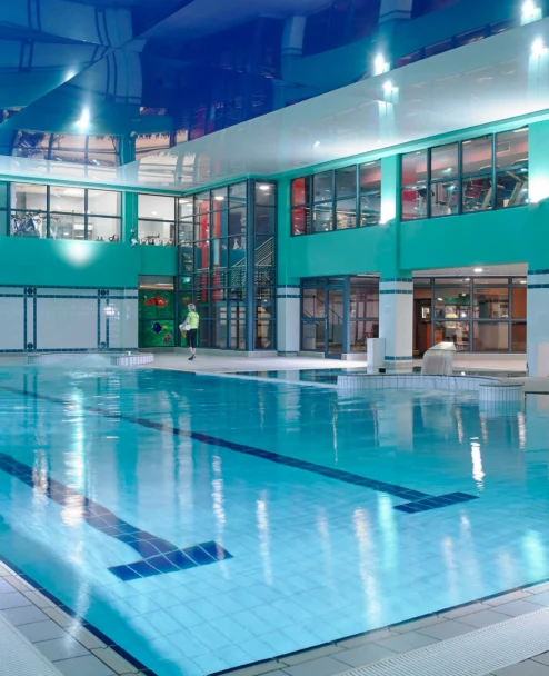 Family enjoying indoor hotel pool area with a parent and child walking by the water, turquoise décor (no visible text)