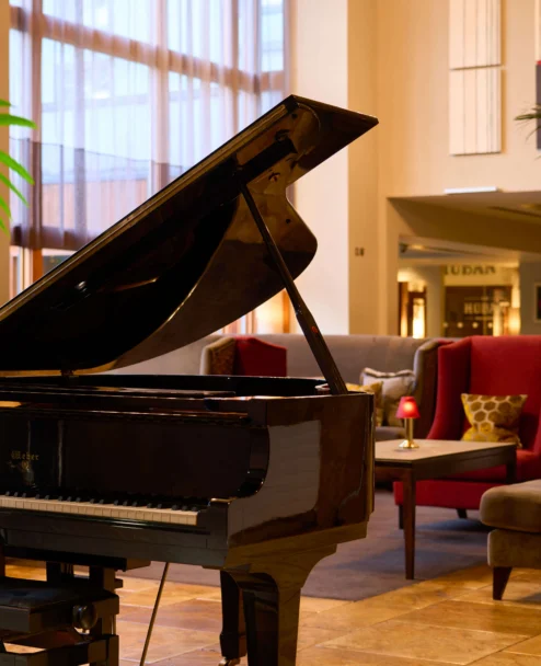 Grand piano in a warm hotel lounge with family seating, soft lamps and plants (no visible text)