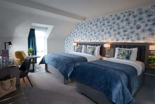 Twin beds in a bright family hotel room with blue floral wallpaper, cosy seating and window curtain