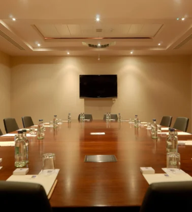 Spacious hotel meeting room with long wooden table, chairs, bottled water and notepads, warm lighting