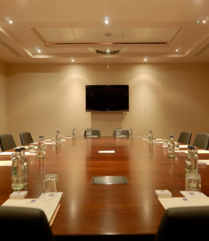 Spacious hotel meeting room with long wooden table, chairs, bottled water and notepads, warm lighting