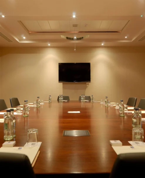 Spacious hotel meeting room with long wooden table, chairs, bottled water and notepads, warm lighting