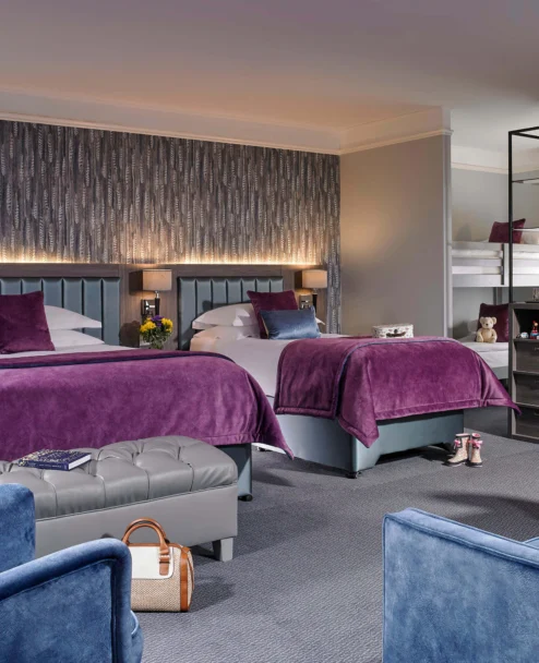 Spacious family hotel room with two double beds, purple throws, seating area and bunk for children