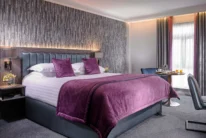 Stylish hotel bedroom with plush purple bedding, bedside lamps, window curtains and breakfast tray