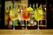 Three colourful cocktails on a bar with lemon, lime and mint garnishes, warm evening setting