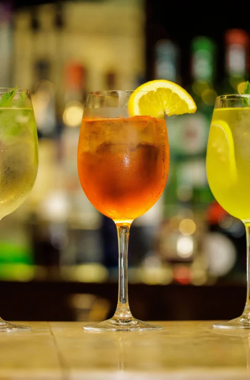 Three colourful cocktails on a bar with lemon, lime and mint garnishes, warm evening setting