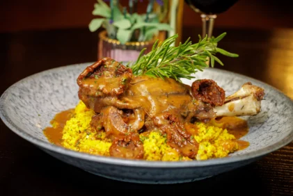 Tender lamb shank with rich gravy and golden couscous, garnished with rosemary on a plate (no visible text)