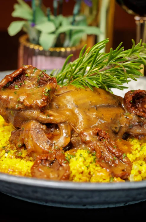 Tender lamb shank with rich gravy and golden couscous, garnished with rosemary on a plate (no visible text)