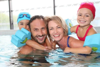 Smiling family of four in a hotel pool with children on parents' backs using float armbands, indoor setting