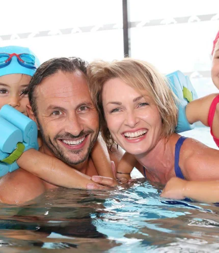 Smiling family of four in a hotel pool with children on parents' backs using float armbands, indoor setting