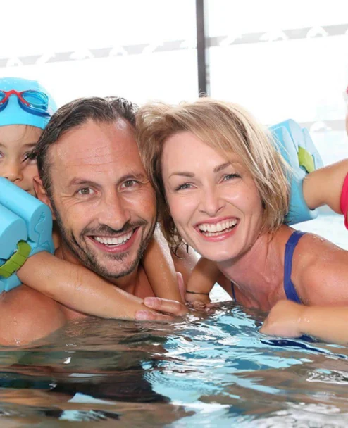 Smiling family of four in a hotel pool with children on parents' backs using float armbands, indoor setting