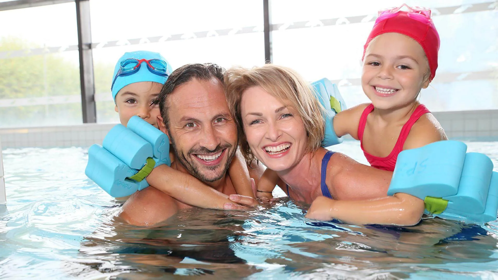 Smiling family of four in a hotel pool with children on parents' backs using float armbands, indoor setting